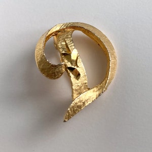 May include: A gold-coloured brooch with a textured surface. The brooch features a flowing, abstract design with a curved shape and a central accent. The metal has a brushed appearance, reflecting light.