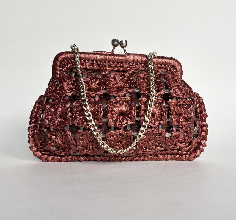 May include: A maroon crocheted clutch with a silver chain strap. The bag features a kiss-lock clasp and a woven pattern. The chain is silver and the bag is a solid maroon color.