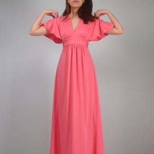 May include: A coral maxi dress with a V-neck and butterfly sleeves. The dress has a fitted waist and a floor-length skirt. The model is wearing the dress. The dress is made of a soft, flowing fabric. The dress is a vibrant coral colour.