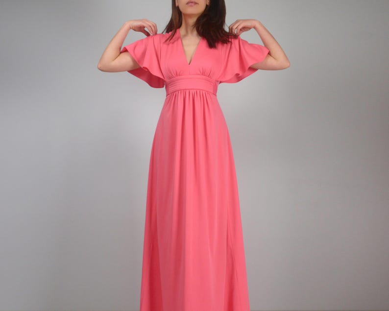 May include: A coral maxi dress with a V-neck and butterfly sleeves. The dress has a fitted waist and a floor-length skirt. The model is wearing the dress. The dress is made of a soft, flowing fabric. The dress is a vibrant coral colour.