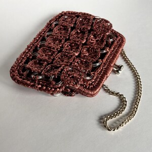 May include: A handmade, rectangular purse in a deep reddish-brown hue. The purse is constructed with a woven pattern and features silver-colored metal rings and a delicate chain strap. The overall design suggests a unique, handcrafted accessory.