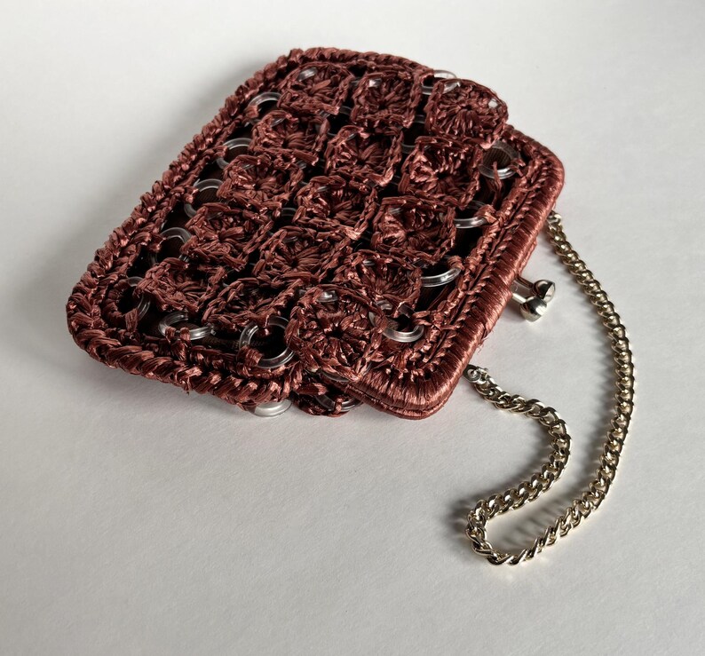May include: A handmade, rectangular purse in a deep reddish-brown hue. The purse is constructed with a woven pattern and features silver-colored metal rings and a delicate chain strap. The overall design suggests a unique, handcrafted accessory.