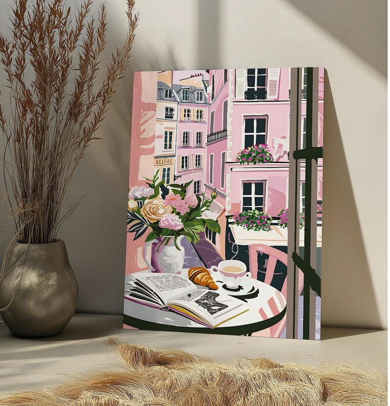 Peut inclure: Une peinture repr&eacute;sentant une sc&egrave;ne de caf&eacute; parisien avec un b&acirc;timent rose, une table avec un livre, un croissant et une tasse de caf&eacute;. Le caf&eacute; est situ&eacute; dans une rue avec d'autres b&acirc;timents en arri&egrave;re-plan. La peinture est encadr&eacute;e et a un passe-partout blanc.