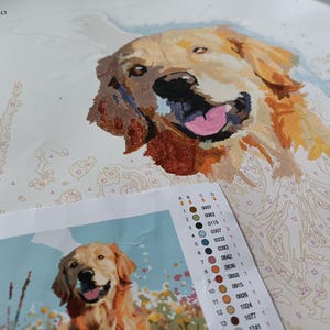 May include: A paint-by-numbers kit featuring a golden retriever dog with its tongue sticking out. The kit includes a numbered canvas, paint pots, and brushes. The numbers on the canvas correspond to the colors of the paint pots.