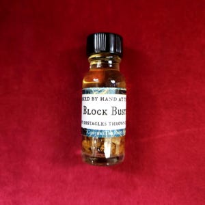 May include: A small glass bottle with a black cap, filled with a light brown liquid and small, solid pieces. The label reads "Block Bust" with additional text. The bottle is set against a red background.