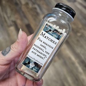 May include: A clear glass bottle filled with white matches, featuring a black lid and a label with text that reads "MATCHES FOR BURNING SAGE, BRIDGES, THE PATRIARCHY, AND MORE!" The label also includes the text "WORKED BY HAND AT THE ALTAR" and "CONJURETHEROOT.COM".