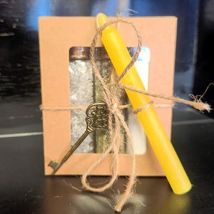 May include: A kraft paper gift box tied with twine, featuring a yellow candle, a decorative key, and two small glass bottles filled with crystals and herbs. The box has a window to view the contents.