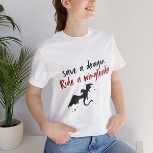 Könnte beinhalten: Weißes T-Shirt mit einer schwarzen Silhouette eines Drachen und dem Text "Save a dragon Ride a wingleader" in roten und schwarzen Buchstaben.