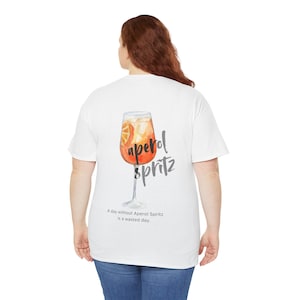 Könnte beinhalten: Weißes T-Shirt mit einer Aquarell-Illustration eines Aperol Spritz Cocktails. Der Text "aperol spritz" ist in schwarzer Kursivschrift geschrieben. Der Text "A day without Aperol Spritz is a wasted day." ist in Schwarz unter der Illustration geschrieben.