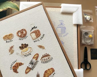 May include: An embroidery kit featuring a framed canvas with stitched illustrations of a bagel, pretzel, white bread, muffin, croissant, baguette, sourdough, cinnamon roll, and challah, each labeled with text, plus accessories including scissors and beads.