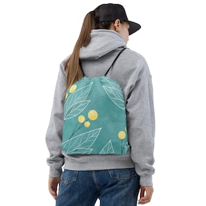 Peut inclure: Un sac à cordon de serrage turquoise avec un motif botanique de feuilles blanches et d'accents circulaires jaunes. Le sac a des cordons noirs et se porte sur les épaules. Le sac est fait d'un matériau doux.