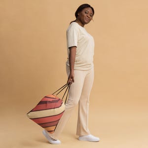 Op de afbeelding: Een persoon houdt een trekkoordtas met horizontale strepen in tinten oranje, rood en crème vast. De tas heeft zwarte trekkoorden. De persoon draagt een crèmekleurig T-shirt en broek, en witte schoenen. De achtergrond is effen beige.