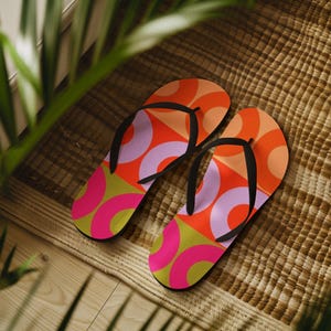May include: A pair of patterned flip-flops with a geometric design. The sandals feature a pattern of orange, pink, light purple, and green shapes. The straps are black. The flip-flops are resting on a woven mat.