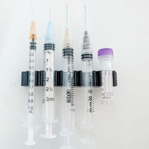 May include: A black plastic holder containing four clear plastic syringes of varying sizes and one small clear vial with a purple cap. The syringes feature printed measurement markings and needle hubs in orange, blue, and white colors.