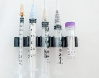 May include: A black plastic holder containing four clear plastic syringes of varying sizes and one small clear vial with a purple cap. The syringes feature printed measurement markings and needle hubs in orange, blue, and white colors.