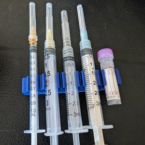 May include: Four clear plastic syringes with varying needle hub colors and a small clear vial with a purple cap, all held in a blue plastic rack. The syringes feature black measurement markings in mL and cc, with capacities ranging from 1 mL to 3 mL.