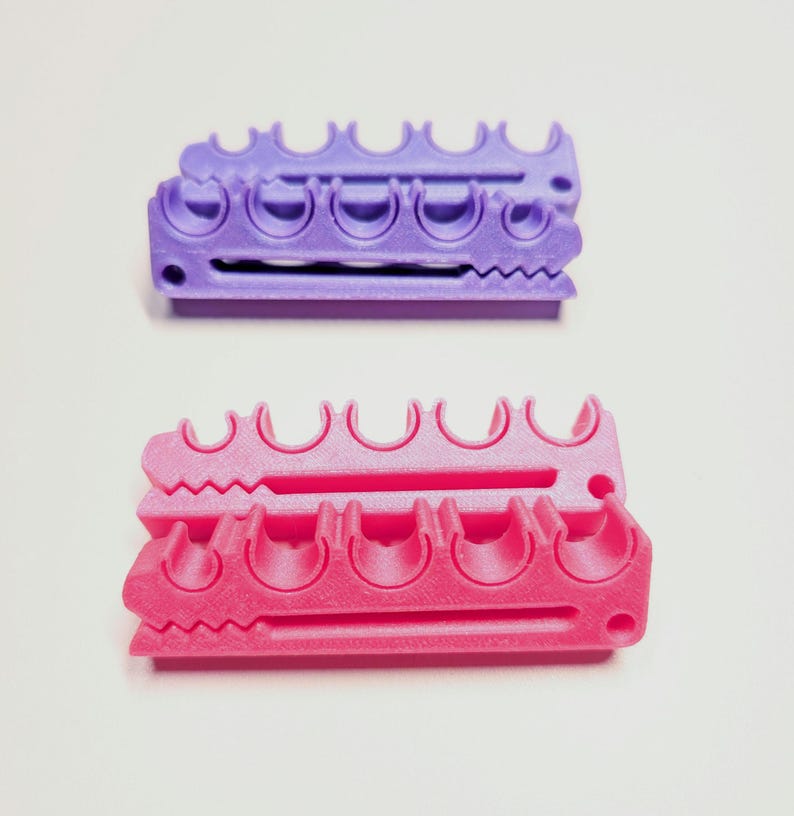 May include: Two plastic organizers, one purple and one pink, each featuring a series of five semi-circular cutouts along the top edge and a long horizontal slot through the center, with a small circular hole at one end of each rectangular unit.