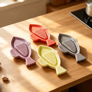 May include: Four colorful fish-shaped ceramic graters in red, lavender, yellow, and gray. Each grater has a textured surface for grating. The graters are arranged on a wooden surface, suggesting a kitchen or dining setting.