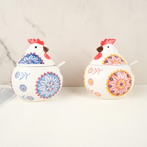 May include: Two white ceramic chicken-shaped jars with lids and small spoons. The jars have red combs, brown beaks, and are decorated with blue, red, and yellow floral patterns. The jars are designed for kitchen use.