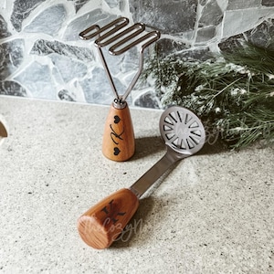 Könnte beinhalten: Zwei Küchenutensilien: ein Kartoffelstampfer und eine Kartoffelpresse. Der Kartoffelstampfer hat einen Kopf aus Edelstahl und einen Holzgriff mit dem Buchstaben "K" und Herzmotiven. Die Kartoffelpresse hat einen kreisförmigen Kopf mit einem Holzgriff, ebenfalls mit dem Buchstaben "K".