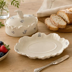Puede incluir: Una mantequillera de cerámica color crema con borde festoneado y un pequeño asa con forma de animal en la tapa. La pieza y la tapa están decoradas con pequeñas flores azules. Un plato ovalado a juego con borde festoneado se sitúa delante.