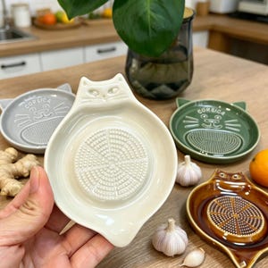 May include: A selection of ceramic cat-shaped graters in cream, grey, green, and brown. Each grater has a textured grating surface and the word "Corsica". The graters are displayed with garlic and ginger.