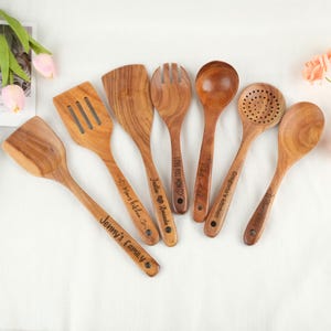 Może przedstawiać: Zestaw siedmiu drewnianych przyborów kuchennych, w tym szpatułka, łyżka szczelinowa i łyżka do serwowania. Każde narzędzie jest grawerowane spersonalizowaną wiadomością. Drewniane łyżki są w kolorze jasnobrązowym i ułożone na białej powierzchni.