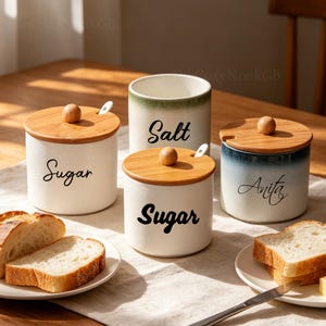 Puede incluir: Cuatro botes de cocina de cerámica con tapas de madera y cucharas pequeñas. Dos botes están etiquetados como "Sugar", uno como "Salt" y otro con el nombre "Anita". Los botes son blancos con un degradado de color en la parte superior.