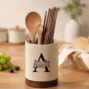 May include: A cylindrical ceramic utensil holder with a speckled beige finish and a dark wood base. The front features a large black letter A with floral accents and the name Alexandra printed in the centre, holding wooden spoons and chopsticks.