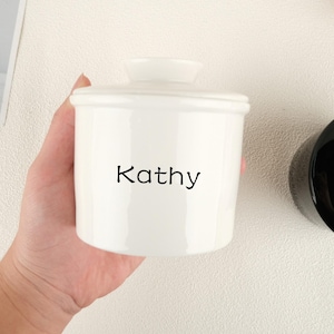 May include: A white ceramic butter dish with a lid, featuring the name "Kathy" in black lettering. The dish has a cylindrical shape and a small handle on the lid. The background is a light, neutral colour.