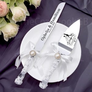 以下が含まれることがあります： レースのハンドルとパールがあしらわれた白いケーキサーバーとナイフのセット。ナイフには「Ophelia & Hamilton March 20, 2025」と刻印されています。サーバーには新郎新婦のシルエットが描かれています。