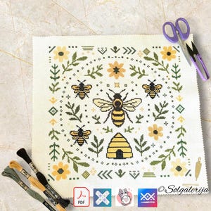 May include: A cross-stitch design with bees, a beehive, and floral motifs in yellow, black, and green on a white canvas. The pattern shows a central bee with wings spread, surrounded by smaller bees and flowers. Purple scissors and embroidery threads are also visible.