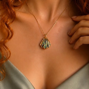 Puede incluir: Un collar dorado con un colgante de concha con acentos iridiscentes verdes y azules. El colgante cuelga de una delicada cadena. El collar lo lleva una persona que lleva una blusa de sat&eacute;n.