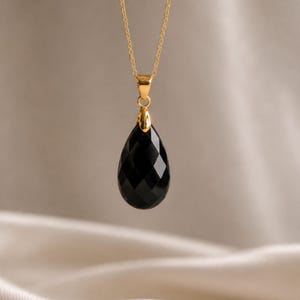 May include: A gold chain necklace with a faceted, teardrop-shaped black pendant. The pendant is suspended from a gold bail and chain, creating a simple yet elegant design. The necklace is displayed against a neutral background.