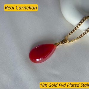 K&ouml;nnte beinhalten: Eine goldfarbene Halskette mit einem tropfenf&ouml;rmigen, leuchtend roten Karneol-Anh&auml;nger. Der Anh&auml;nger ist glatt und poliert. Der Text "Real Carnelian" und "18K Gold Pvd Plated Stainless Steel" sind im Bild.