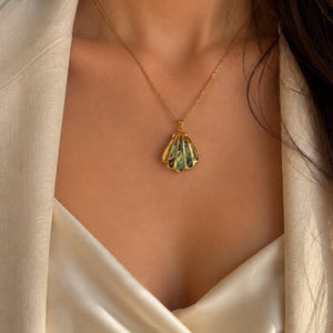 Puede incluir: Un collar con colgante de concha dorada con una cadena dorada. El colgante tiene una incrustaci&oacute;n iridiscente verde y azul. El collar se lleva sobre una blusa de sat&eacute;n color crema y una blazer beige.