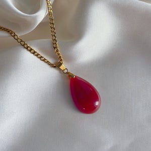 K&ouml;nnte beinhalten: Eine goldfarbene Halskette mit einem tropfenf&ouml;rmigen, tiefrosa Anh&auml;nger. Der Anh&auml;nger ist glatt und gl&auml;nzend. Die Halskette liegt auf einem wei&szlig;en, seidigen Stoff.
