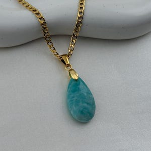 May include: Gold-toned chain necklace with a teardrop-shaped pendant. The pendant is a light blue-green stone with a marbled appearance. The necklace is displayed on a white surface.
