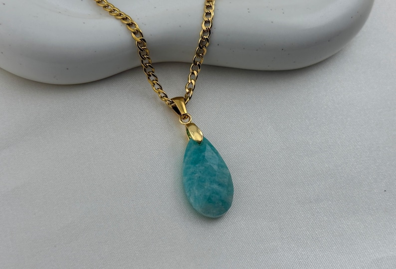 May include: Gold-toned chain necklace with a teardrop-shaped pendant. The pendant is a light blue-green stone with a marbled appearance. The necklace is displayed on a white surface.