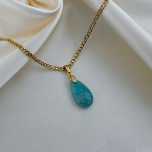 May include: A gold-colored chain necklace with a teardrop-shaped, faceted, turquoise pendant. The pendant is suspended from a small gold-colored bail. The necklace rests on a white, silky fabric.