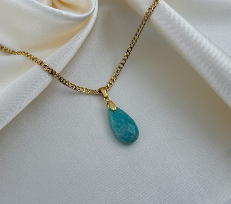 May include: A gold-colored chain necklace with a teardrop-shaped, faceted, turquoise pendant. The pendant is suspended from a small gold-colored bail. The necklace rests on a white, silky fabric.