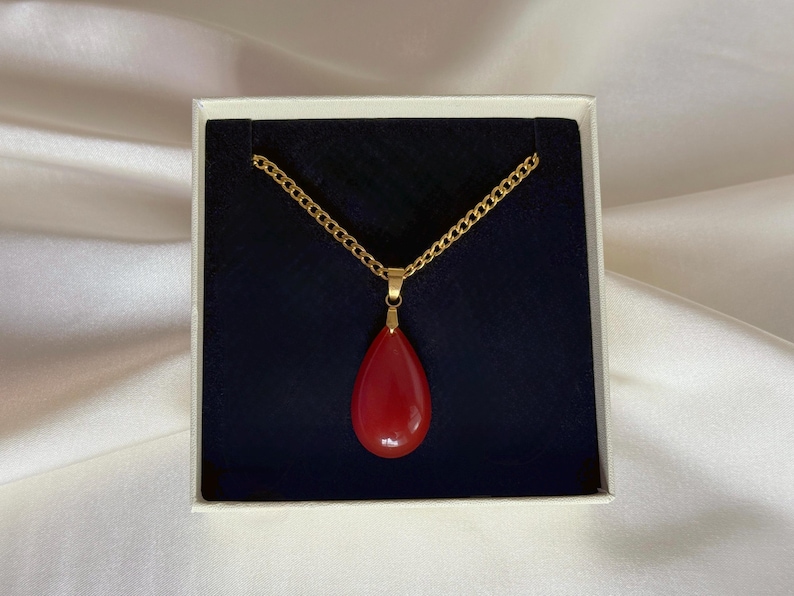 May include: A gold-colored chain necklace with a teardrop-shaped, red pendant. The necklace is presented in a white box with a dark blue interior. The pendant is smooth and glossy.