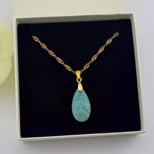 May include: A gold-colored chain necklace with a teardrop-shaped, light blue pendant. The necklace is displayed in a white box with a black interior. A yellow flower is partially visible in the background.