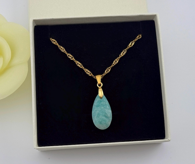 May include: A gold-colored chain necklace with a teardrop-shaped, light blue pendant. The necklace is displayed in a white box with a black interior. A yellow flower is partially visible in the background.