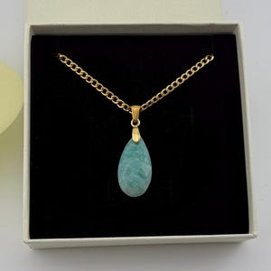 May include: A gold-colored chain necklace with a teardrop-shaped, light blue pendant. The necklace is displayed in a white gift box with a black interior. A yellow rose is visible in the background.
