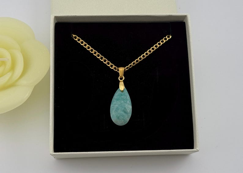 May include: A gold-colored chain necklace with a teardrop-shaped, light blue pendant. The necklace is displayed in a white gift box with a black interior. A yellow rose is visible in the background.
