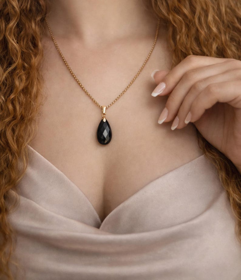 May include: A gold-colored necklace with a teardrop-shaped black pendant. The pendant is faceted, catching the light. The necklace is worn against a light pink fabric.