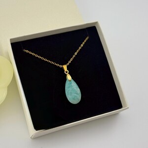 May include: A gold-colored chain necklace with a teardrop-shaped, light blue pendant. The necklace is displayed inside a white gift box with a black interior. A yellow flower is visible in the background.