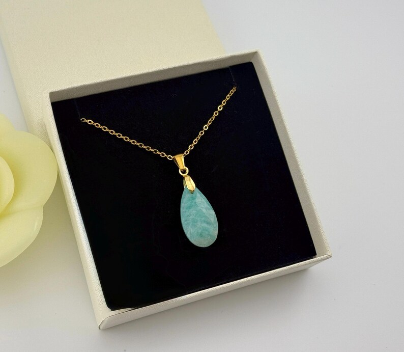 May include: A gold-colored chain necklace with a teardrop-shaped, light blue pendant. The necklace is displayed inside a white gift box with a black interior. A yellow flower is visible in the background.