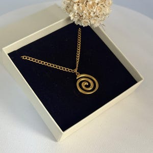 May include: A gold-colored spiral pendant necklace displayed in an open, cream-colored gift box with a black velvet interior. The necklace chain is also gold-colored. A small bouquet of dried flowers sits above the box.